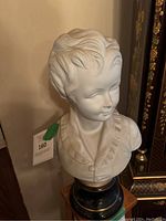 Front view of large bisque porcelain bust of a child with detailed hair and period clothing, resting on a decorative base.