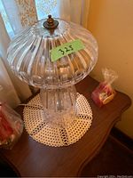 Photo of one lamp showing rounded ribbed glass shade, brass finial on top, clear vertical fluted base, sitting on a wooden table with a crocheted doily underneath
