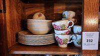 Photo of stacked white plates, bowls, and teacups with rose floral decorations, stored on wooden shelf.