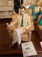 Photo shows a detailed resin figurine by Vittorio Tessaro of a man sitting on a tan couch, holding an object, dressed in patterned blazer and teal tie.