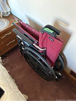 Folded Magic brand wheelchair leaning against a wall near furniture, showing maroon fabric and black large rear wheels.