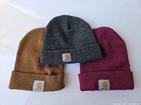 Three Carhartt winter knit hats in brown, gray, and red arranged side by side on white background, showing front logo patches