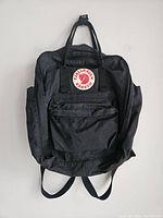 Full black Fjallraven Kanken backpack showing front, shoulder straps, carrying handles, front pocket, and side pockets.