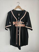 Front view of black Harley Davidson jersey shirt with white buttons and orange/white trim along edges. "HARLEY DAVIDSON" text on chest and branding on sleeves.