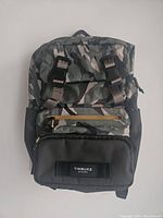 Front view of Timbuk2 Curator Backpack in camouflage pattern showing main compartment with two black buckles, front zippered pocket with Timbuk2 label, and black bottom panel.