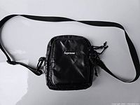 Full view of the black Supreme crossbody bag with adjustable shoulder strap laid flat on a white surface.