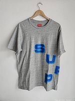 Front view of grey Supreme t-shirt showing diagonal blue letters spelling 'SUP'