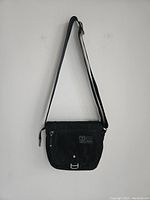 Full view of black TUMI Tech shoulder bag hanging against white wall, showing flap with zippered pocket and adjustable strap.