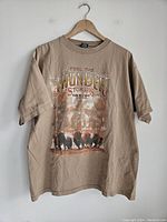 Full view of tan T-shirt with front graphic showing motorcycles and text 'Feel The Thunder Sturgis Black Hills Rally 2003'. Shirt is on a hanger against a white wall.