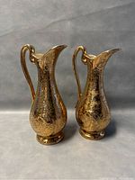Pair of gold embossed ceramic 12-inch ewer pitchers, showing front and side views with handles and elongated spouts.