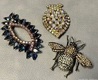 Three vintage costume brooches laid out on a grey textured surface: a bee with crystals, a shield-shaped piece with iridescent crystals, and an elongated oval piece with dark blue crystals.