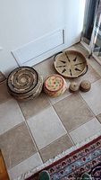 Five baskets grouped on tile floor near vent and door. Includes large sunburst patterned basket and other smaller lidded and open baskets.