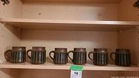 Six brown ceramic coffee mugs with green vertical stripes displayed on a shelf.