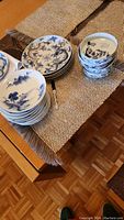 Three stacks of ceramic plates and bowls displayed on a woven table runner over a wooden table. Blue and multicolored painted designs visible.