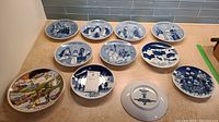 Ten blue and white collector Christmas plates and one colorful Norge souvenir plate, arranged on kitchen counter.