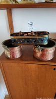 Two round copper pots with metal handles and an oval copper planter with star cutout design on top of a wood cabinet.