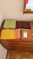 Photo showing stacks of vintage linen table napkins in various colors including green, purple, brown, yellow, and peach with some having fringed edges, placed on top of a wooden chest of drawers.