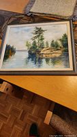 Framed oil painting showing a landscape with a lake, pine trees, and rocky shore.