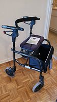 Side front angle view of Dana Douglas Nexus 3 rollator walker with padded seat, hand brakes, and under-seat pouch. User manual on seat.