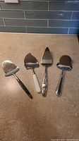 Four metal serving utensils laid out including one pewter cake server and three cheese slicers with different handles.