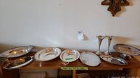 Wide shot showing multiple silver plated trays, vases, and cake stand on wooden surfaces against a white wall