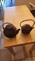 Two brown ceramic teapots with woven wicker handles placed on a wooden table, showing size difference.