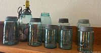 Eight blue Ball Mason jars, one converted to a lamp with electrical fitting, arranged on a wooden surface against a wall.