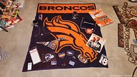 Overview of all Broncos branded items arranged on the orange and navy throw blanket featuring a large Bronco logo and 'BRONCOS' text.