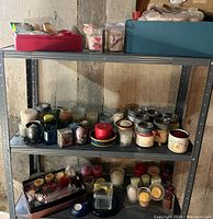 Metal shelving unit with multiple jar candles, votives, and candle holders arranged on middle and bottom shelves, and trays of smaller candles.