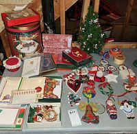 Photo showing assortment of Christmas decorations, holiday cards, large decorative tin, small tabletop Christmas tree, and various ornaments laid out on a table.