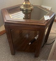 Hexagon vintage side table shown with a glass inset top and wooden frame. The table is placed on carpeted floor with a brass lamp on top.