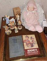 Overview image showing porcelain baby doll seated on lace chair surrounded by five Hummel figurines and two vintage cookbooks on the table.