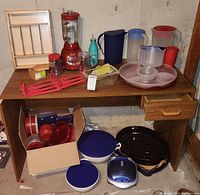 Full view of a wooden desk with a red vintage blender, assorted plastic pitchers, plastic serving tray with compartments, glass Pyrex loaf pan with lid, small glass jars, a wooden rack, and a cardboard box with additional plastic storage containers below desk along with enamel roasting pans.
