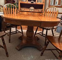 Front-facing image of the oak oval dining table with one leaf inserted, surrounded by 4 of the 6 chairs, showing the wood grain and central pedestal base.