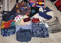 Overview photo showing 13 assorted bags and purses arranged on a floor rug, featuring various colors and styles such as quilted floral patterns, solid colored leather, and fabric bags.
