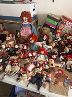 Full view of a table with numerous Raggedy Ann and Andy dolls, a teddy bear, and boxed items in the background.