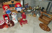 Overview of the assortment including Raggedy Ann & Andy dolls on the bench, snowman plush, doll heads, red containers, doll in red dress, wooden caddy, and numerous rocking horse figurines in the background.