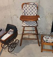 Full view of vintage wooden high chair with tray, antique doll pram to the left, and child's stool to the right, situated against concrete wall.