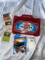 Photo shows Bakugan Battle Brawlers carrying case, three Pokémon cards, and Hot Wheels car in original packaging on white fabric background.