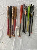Top view of all eight bats and autographed baseball arranged on white cloth background. Shows wood and composite bats with grips and colors.