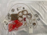 Top view of assorted costume jewelry spread on white cloth showing necklaces, bracelets, rings, earrings, and red organza bags with pearl-like bead necklaces.