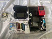 All items arranged on white surface: PSP in case, stylus pack, Game Boy Color, 3DS in case, two keychains in packaging, and game cartridge