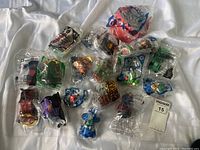 Photo showing multiple unopened McDonald's Happy Meal toys including Hot Wheels, Mario Kart, Super Mario, and superhero figures all individually sealed in clear plastic bags on a white fabric background.