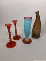 Photo of a blue blown glass vase with pink rim, yellow base and black dots, a brown ribbed glass vase, and a pair of matte orange twisted glass candleholders.