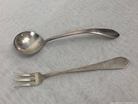 Photo of the sterling silver jelly serving spoon and cocktail fork laid out on a light patterned fabric surface, showing the length and shape of each piece.