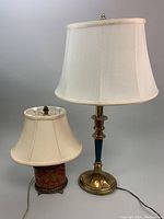 Two vintage table lamps side by side on a neutral background: a taller brass and ceramic lamp with a large cream fabric shade, and a shorter red and gold porcelain lamp with a smaller cream fabric shade.