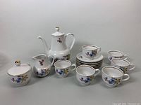 Full view of the tea set showing teapot, creamer, sugar bowl, six cups, and six saucers all in white porcelain with floral pattern and gold accents.