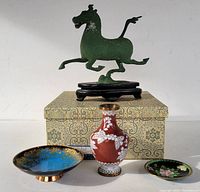 The lot arranged with the Tang Dynasty-style metal horse figurine mounted on a black wooden stand, a small cloisonné vase with red and white floral design, a small cloisonné dish with green and floral pattern, and a blue and gold art enamel trinket tray, all displayed on a gift box with patterned fabric.