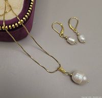 Close-up view of gold overlay necklace with freshwater pearl pendant and matching pearl earrings next to the purple velvet zipper box.