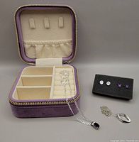 Zippered purple velvet jewelry storage box open with compartments, two silver necklaces laid on ring slots, earrings on black foam nearby.